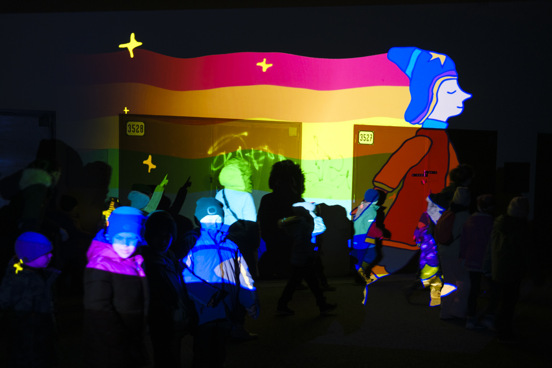 Photo of a video projection animation on a wall while kids try touching the figure. A mage walking with a rainbow